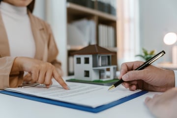 Un cliente firma un documento junto a una maqueta de casa mientras una asesora le señala la página