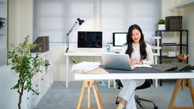Una mujer sonriente toma notas en un escritorio con una laptop y pilas de documentos, en una oficina luminosa