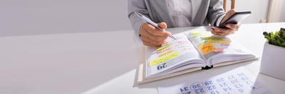 Una persona escribe en una agenda llena de notas adhesivas de colores junto a un calendario de escritorio