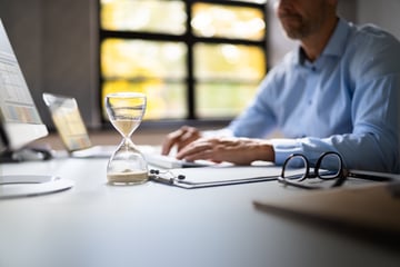 Un reloj de arena sobre un escritorio, frente a un hombre que escribe en el teclado de una computadora