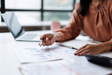 Person reviewing financial reports and charts with a pen, next to a laptop and a calculator