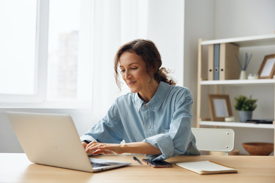Smiling woman in a denim shirt working on a laptop at a desk in a bright home office