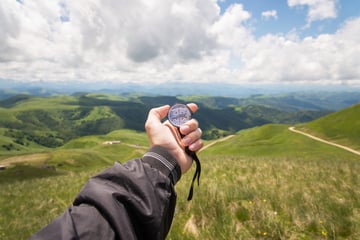 Hand holding a compass in front of green mountain hills under a cloudy sky